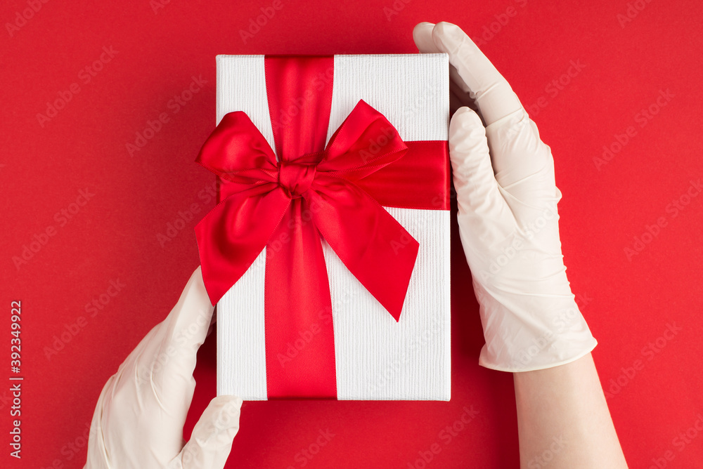 Holiday celebration during quarantine concept. Top above overhead view photo of female hands in white gloves holding white giftbox with red ribbon isolated on red background