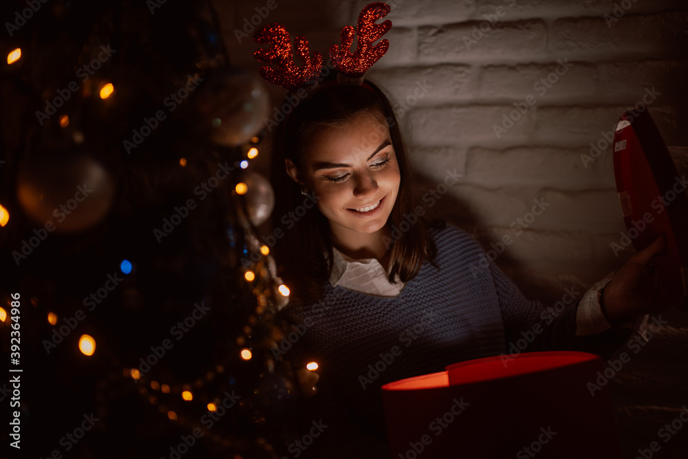 A young smiling teenage girl with horns on her head looks at a red gift box with a ribbon, while in the background is a Christmas tree. Opening of presents late on Christmas Eve