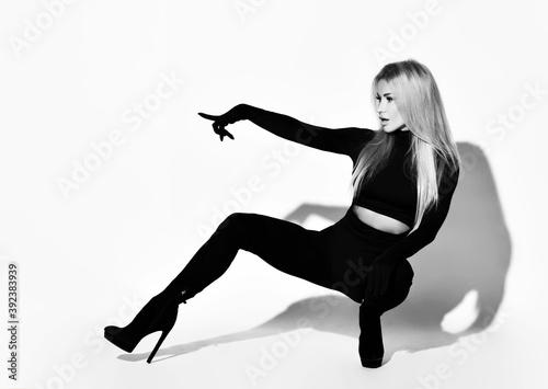 Slim blonde woman with pouty lips in tight black overall suit, high-heeled shoes and gloves sits squatted pointing finger aside at free copy space. Black and white full-growth portrait