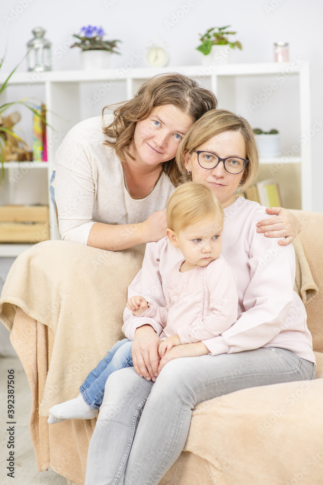Two young women are sitting in the living room on the sofa with a child and smiling to their daughter. Gay lesbian couple at home with a baby. Lesbian family