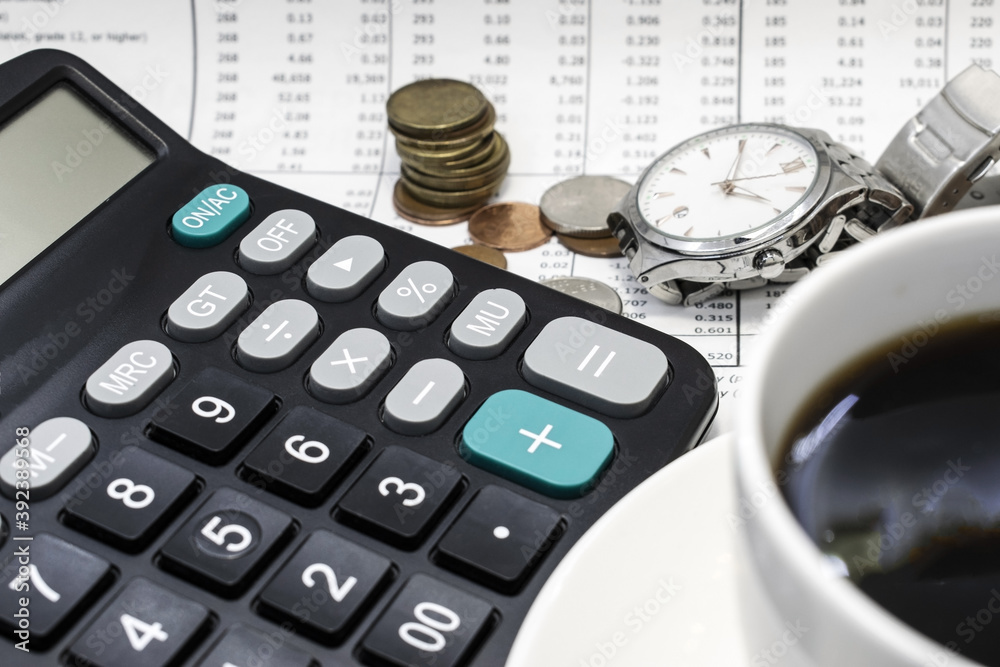 The black calculator is on the blurry background of a document with coins a watch and a Cup of coffee. Business and financial concept The black calculator is on the blurry background of a document with coins a watch and a Cup of coffee. Business and financial concept