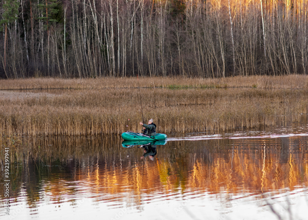 Opposite bank of lake in the autumn, fisherman with a boat in the lake, colorful tree silhouettes and reflections in the water, Latvian landscape Opposite bank of lake in the autumn, fisherman with a boat in the lake, colorful tree silhouettes and reflections in the water, Latvian landscape