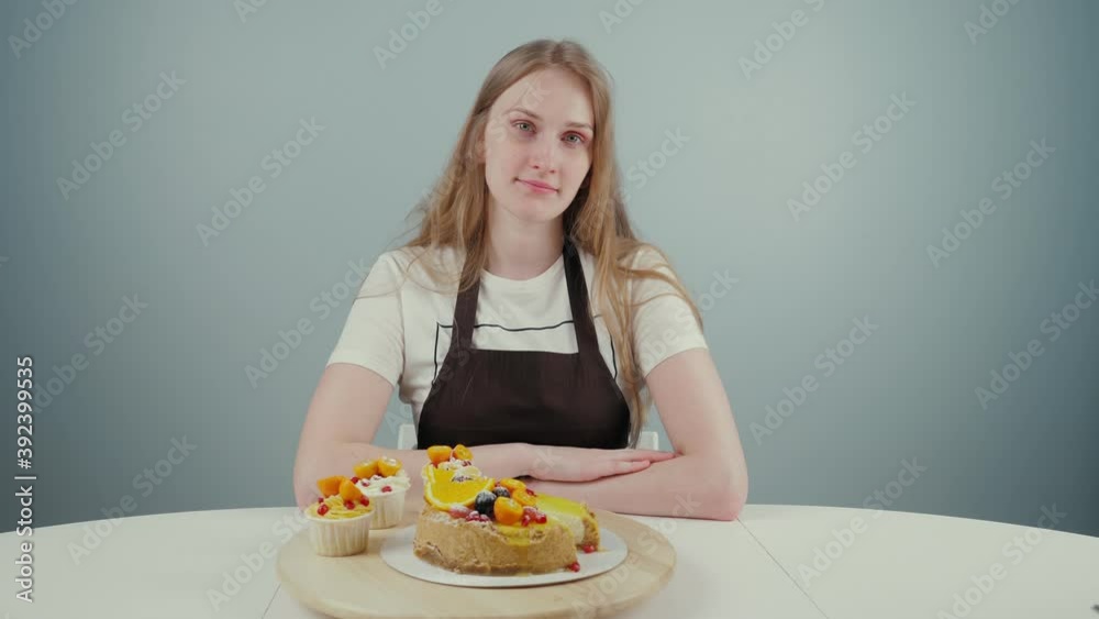 A young girl made cake and cupcakes. Shows the result of her work. Long-haired lady twirls sweets on the table, then looks into the camera. Neutral background. Medium shot. A young girl made cake and cupcakes. Shows the result of her work. Long-haired lady twirls sweets on the table, then looks into the camera. Neutral background. Medium shot.