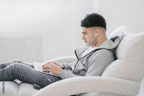 a young man is working at home on a laptop computer, doing an Economics assignment on his new project. He is dressed in comfortable gray clothing and has wireless headphones in his ears