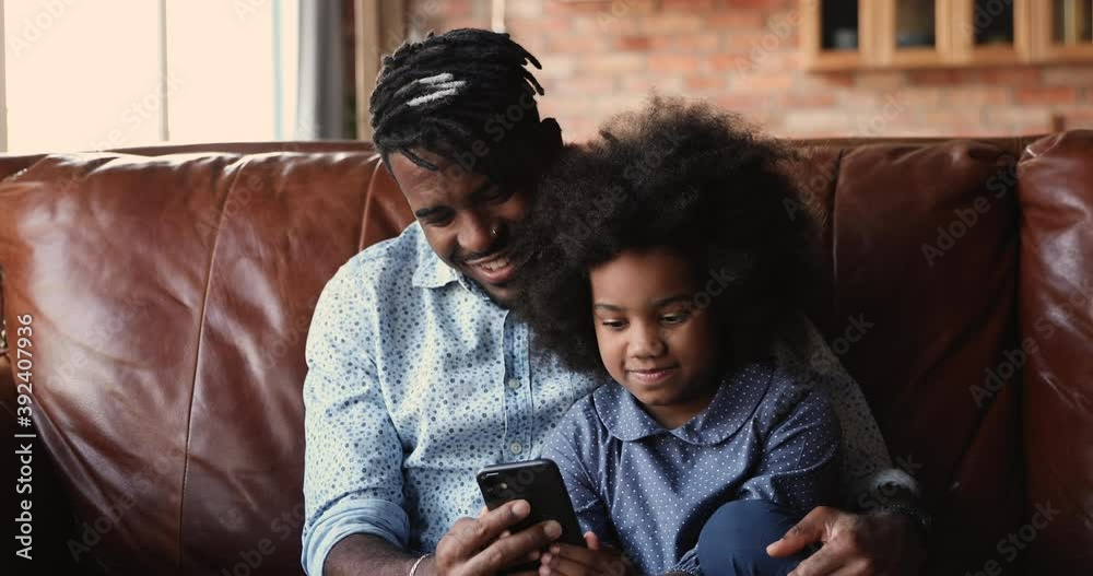 Loving african dad spend time with daughter sit on couch having fun use smartphone and internet. Play games, enjoy new cool app. Parental control, modern tech usage, device overuse, lifestyle concept Loving african dad spend time with daughter sit on couch having fun use smartphone and internet. Play games, enjoy new cool app. Parental control, modern tech usage, device overuse, lifestyle concept