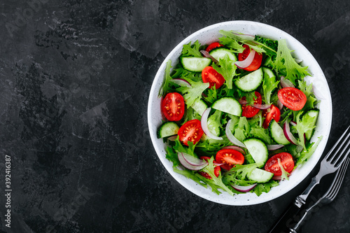 Healthy green salad with fresh tomato, cucumber, red onion and lettuce in bowl on dark stone background.