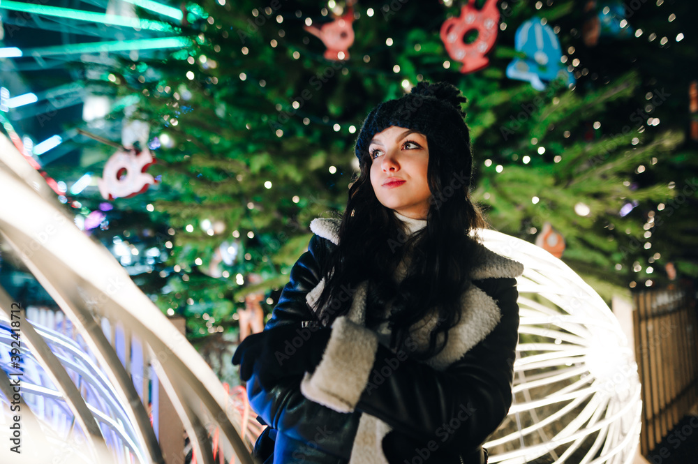 Attractive girl stands in the evening near the Christmas tree and Christmas decorations and looks away with a pensive face, wearing warm clothes. New Year and Christmas Attractive girl stands in the evening near the Christmas tree and Christmas decorations and looks away with a pensive face, wearing warm clothes. New Year and Christmas