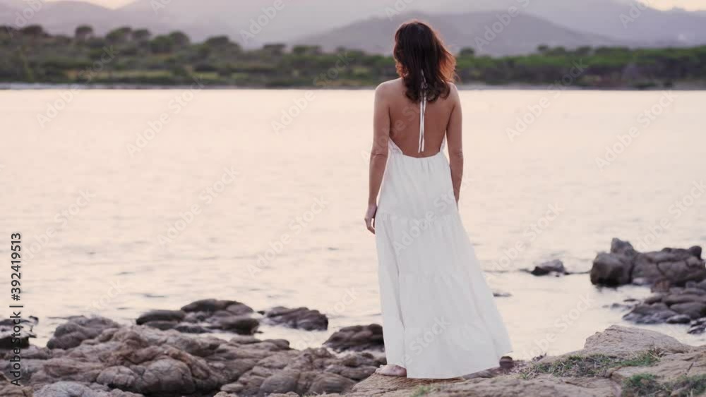 Fantasy scene of unrecognizable Caucasian woman standing on the rocks near the water looking at the sun goes down at the horizon - Back view of pensive female person wearing long dress in barefoot Fantasy scene of unrecognizable Caucasian woman standing on the rocks near the water looking at the sun goes down at the horizon - Back view of pensive female person wearing long dress in barefoot
