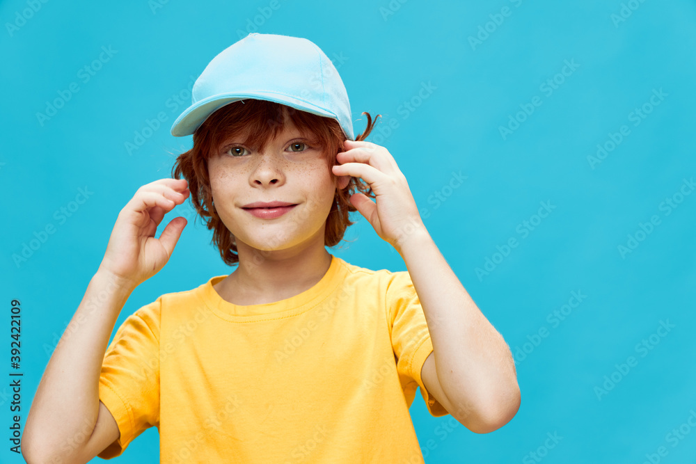 red-haired boy with a blue cap on his head holds his hands near his face yellow t-shirt 