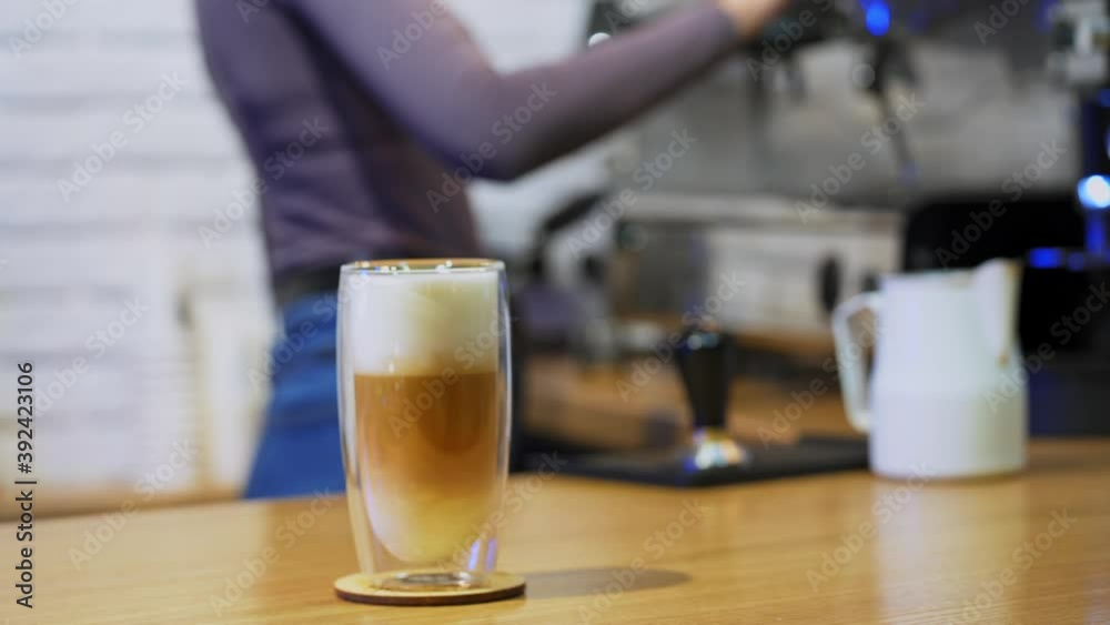 Cappuccino or latte in glass. Woman puts drink from coffee machine. Fresh coffee with whipped cream in tall clear glass on table. Close-up. Cappuccino or latte in glass. Woman puts drink from coffee machine. Fresh coffee with whipped cream in tall clear glass on table. Close-up.