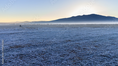 Paisaje azul con suelo congelado por el frío invernal y amanecer dorado en el horizonte. Montaña helada con niebla al fondo.