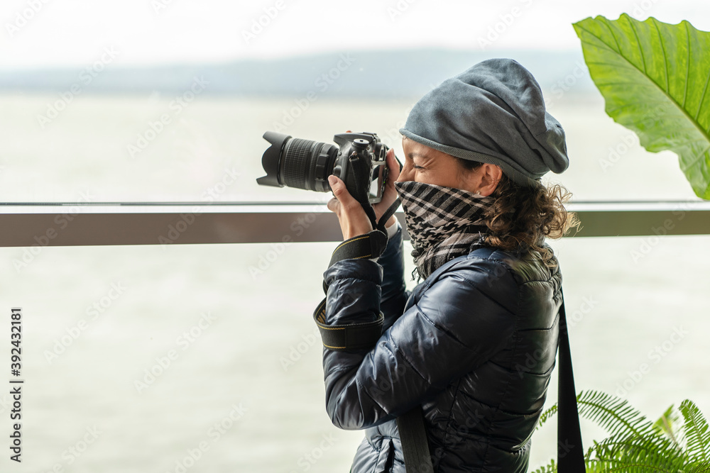 Obraz premium Young female photographer with bandana standing near large window and taking picture of river
