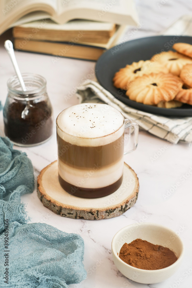 An angle view of a cup of layered macchiato coffee topped with foam with books and plate of cookies in background. A drink is based on the espresso and steamed milk combination. It's similar to latte