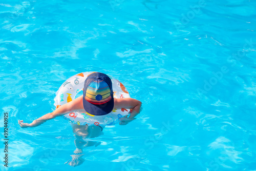 A child in a large colored baseball cap swims in the clear blue water of a pool with a children's inflatable circle