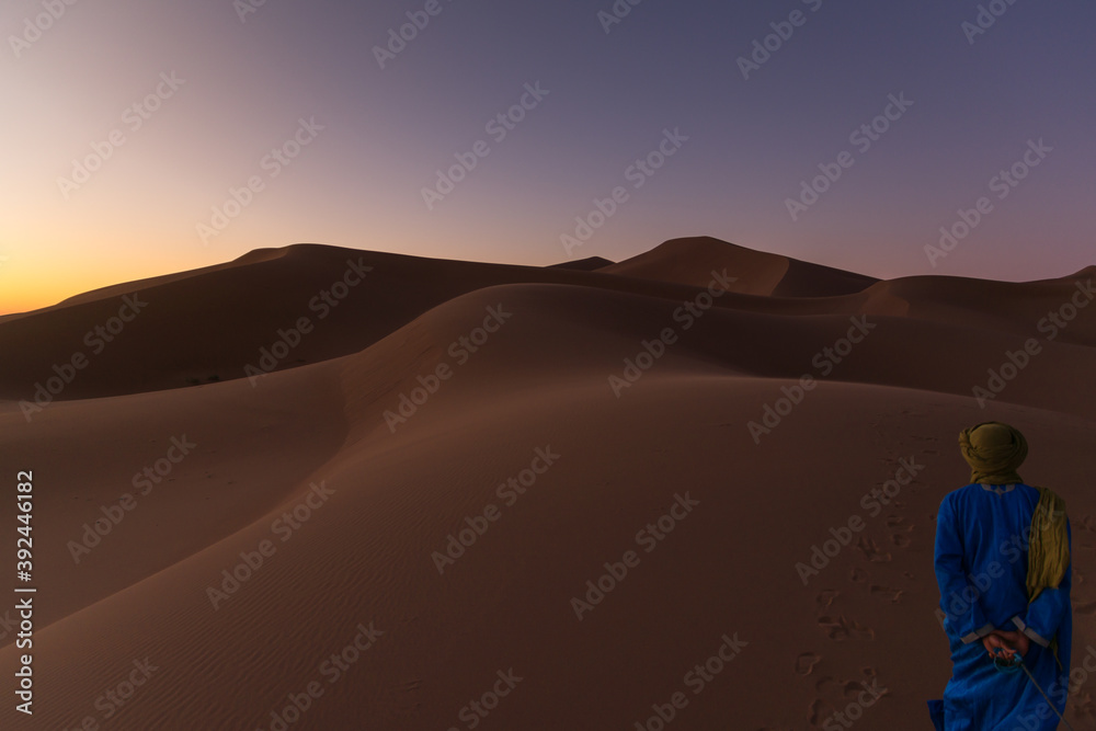 Unrecognizable Berber man walking on a dreamy desert at Twilight of dawn. Desert dune of Erg Chigaga, at the gates of the Sahara. Morocco. Concept of travel and adventure.