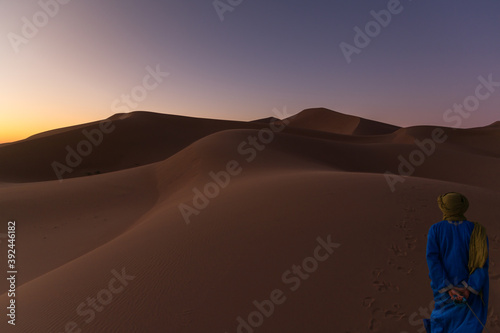 Unrecognizable Berber man walking on a dreamy desert at Twilight of dawn. Desert dune of Erg Chigaga  at the gates of the Sahara. Morocco. Concept of travel and adventure.