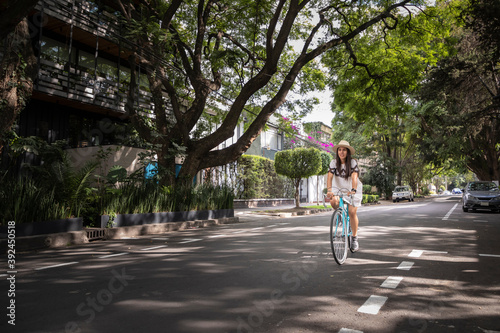 Mujer paseando en bicicleta en la calle con arboles a los lados. Mujer usando vestido blanco y sombrero mientras pasea en bici.