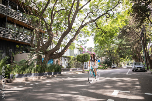 Mujer paseando en bicicleta en la calle con arboles a los lados. Mujer usando vestido blanco y sombrero mientras pasea en bici.