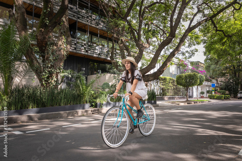 Mujer paseando en bicicleta en la calle con arboles a los lados. Mujer usando vestido blanco y sombrero mientras pasea en bici. © Edgar1 BJ