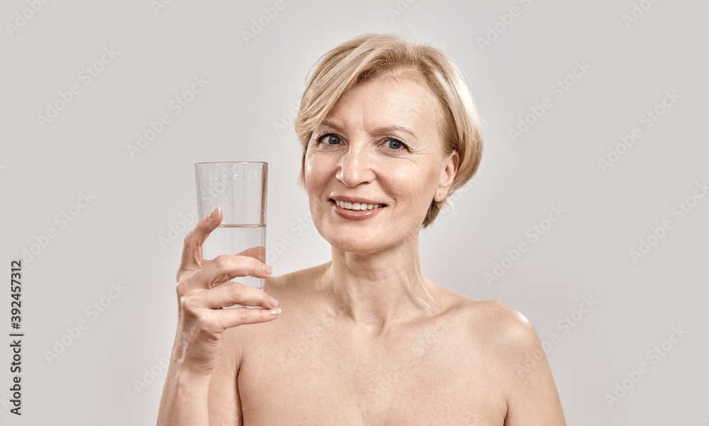 Portrait of attractive middle aged woman smiling at camera while holding glass of water in her hand, posing isolated over grey background Portrait of attractive middle aged woman smiling at camera while holding glass of water in her hand, posing isolated over grey background