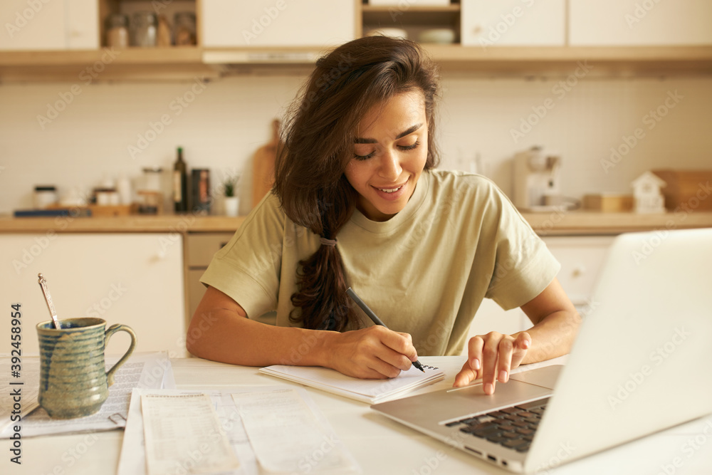 Indoor image of cute Arabic student girl in t-shirt sitting at dining table in front of open laptop, studying distantly, drinking morning coffee and making notes in copybook, listening to lecture Indoor image of cute Arabic student girl in t-shirt sitting at dining table in front of open laptop, studying distantly, drinking morning coffee and making notes in copybook, listening to lecture