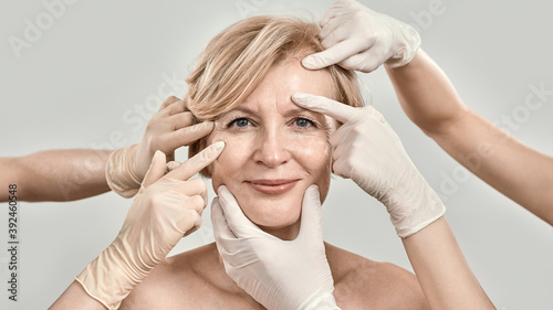 Beauty portrait of middle aged woman smiling at camera. Beautician hands in gloves checking female face skin isolated against grey background