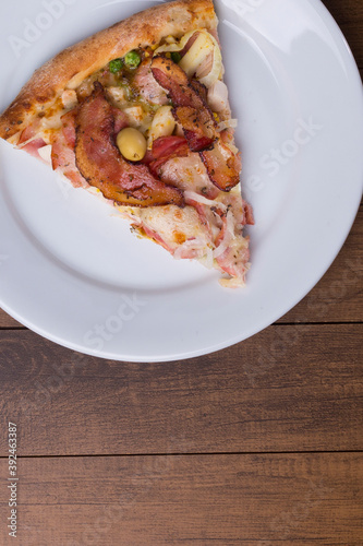 Pizza slice served on the plate. Portuguese pizza made with ham, pea egg, heart of palm, pepperoni, onion and mozzarella and bacon. Close-up photo with bottom space for texts.