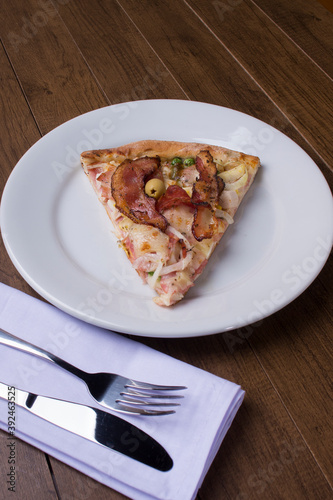 Pizza slice served on the plate. Portuguese pizza made with ham, pea egg, heart of palm, pepperoni, onion and mozzarella and bacon. Fork and knife on a napkin. Centralized elements.