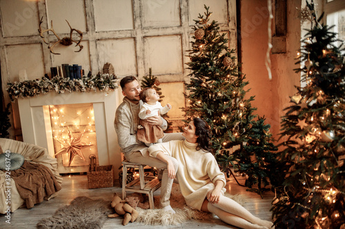Wonderful parents with little daughter sit in living room near the fireplace and Christmas tree, cuddling, enjoy cozy winter holidays together, new year celebration concept