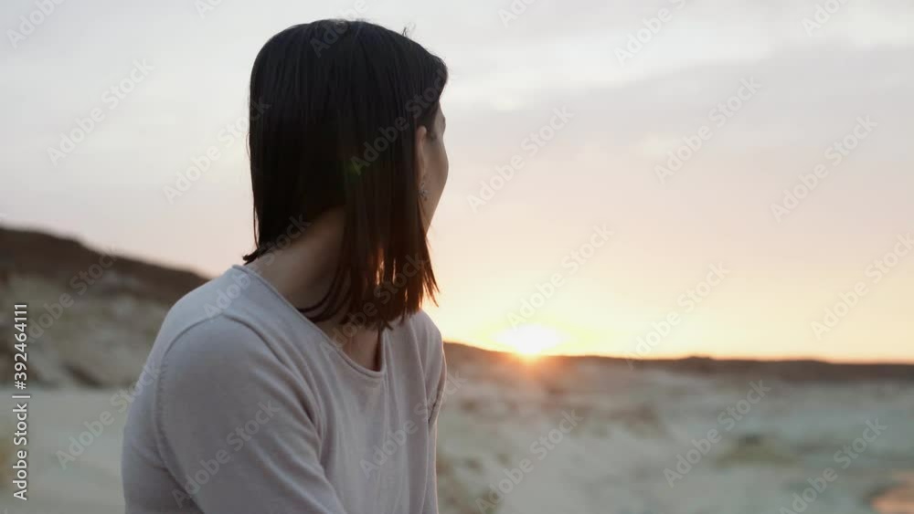 Close up of young brunette in blue skirt sits on top of sand hill with sunset on background Close up of young brunette in blue skirt sits on top of sand hill with sunset on background