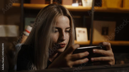 Young blond beautiful woman stretching on the sofa is playing games with smartphone and rejoicing