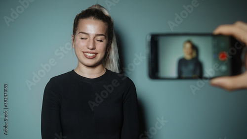 Young blonde beautiful attractive woman is recording video taking vlog with phone in cyan-color studio background