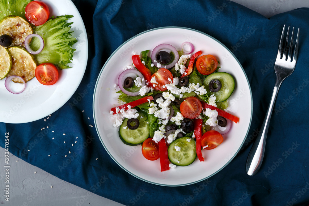 White plate with grilled zucchini with cherry tomatoes on lettuce leaves and Greek salad on a blue napkin with a fork. White plate with grilled zucchini with cherry tomatoes on lettuce leaves and Greek salad on a blue napkin with a fork.