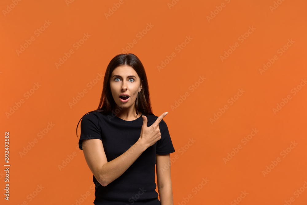 Surprised caucasian woman in black t-shirt with stupefied expression, points with index finger at upper right corner, shows incredible thing, opens mouth. Isolated over an orange background Surprised caucasian woman in black t-shirt with stupefied expression, points with index finger at upper right corner, shows incredible thing, opens mouth. Isolated over an orange background