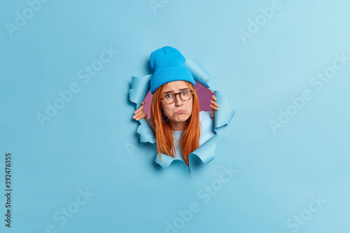 Disappointed upset young woman purses lips with offensive expression looks sadly at camera has long red hair wears hat and transparent glasses sticks head out of ripped hole blue paper wall.