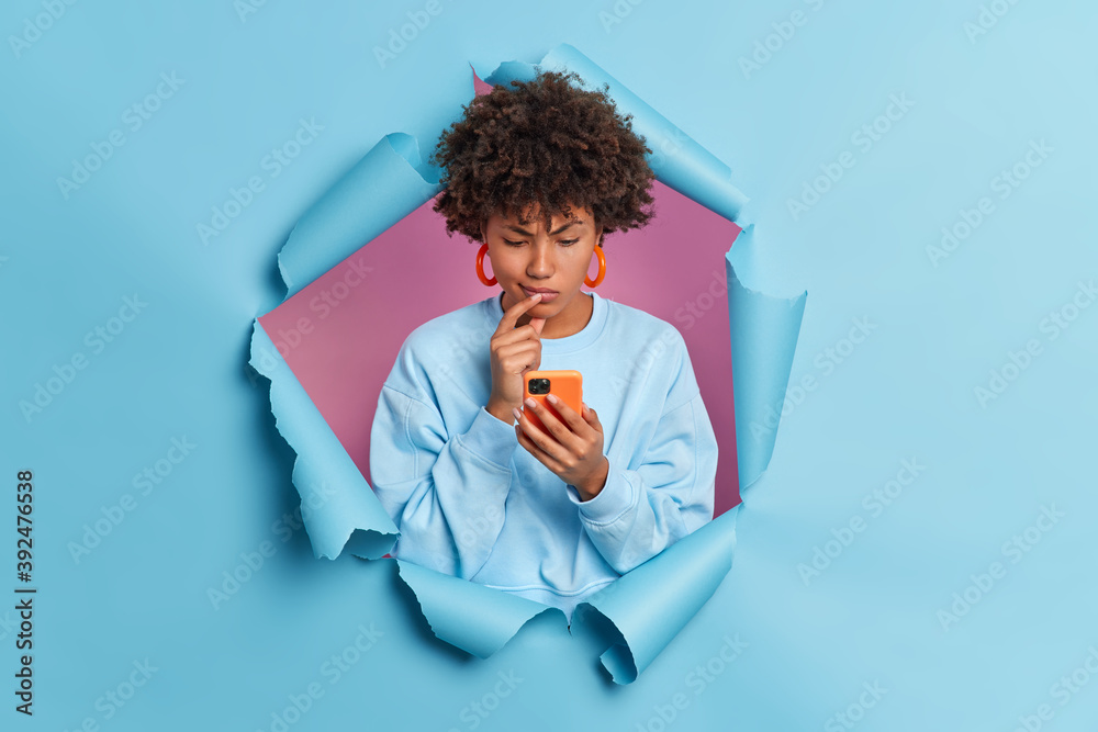Puzzled dark skinned young Afro American woman looks doubtful concentrated in smartphone display reads comments under post on website wears casual jumper stands in studio through torn paper hole - obrazy, fototapety, plakaty 