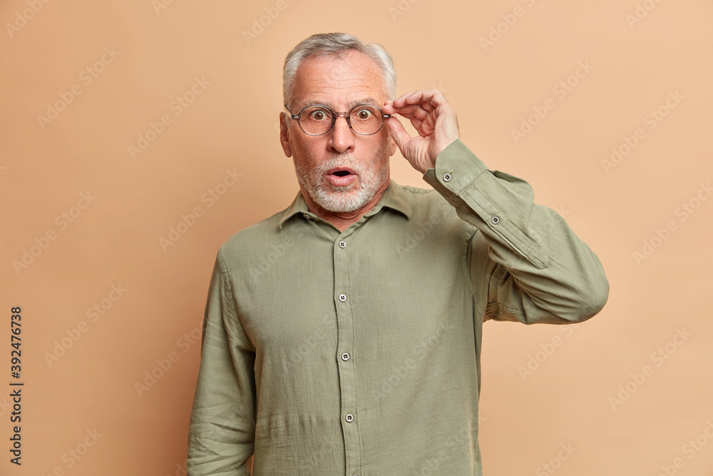 Shocked handsome stunned bearded elderly man has grey hair opens mouth widely keeps hand on spectacles cannot believe in shocking news wears formal shirt poses against brown studio background.