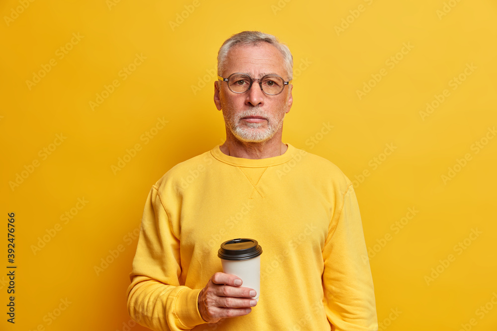 Portrait of bearded grey haired man holds takeaway coffee cup dressed in casual jumper looks directly at camera isolated over yellow background satisfied with free time. People leisure drinks Portrait of bearded grey haired man holds takeaway coffee cup dressed in casual jumper looks directly at camera isolated over yellow background satisfied with free time. People leisure drinks