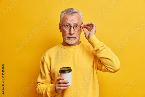 Photo of surprised bearded mature man keeps hand on spectacles drinks takeaway coffee hears astonishing news wears casual jumper isolated over vivid yellow background. People lifestyle concept © wayhome.studio 
