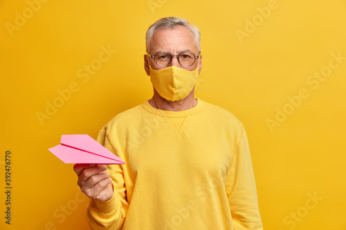 Serious grey haired man looks directly at camera wears transparent glasses protective mask and holds paper airplane dressed in casual yellow jumper being infected with coronavirus. Age and health care