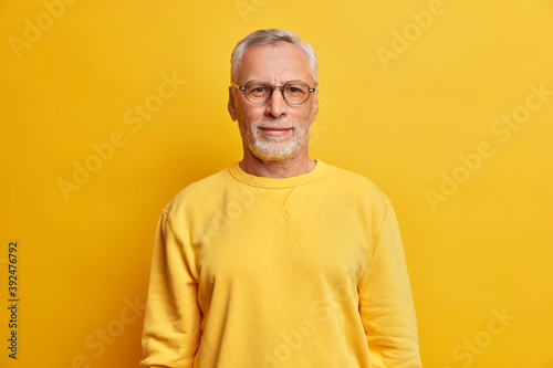 Horizontal shot of grey haired man with wrinkles wears glasses and casual yellow jumper looks directly at camera has satisfied expression poses indoor. Grandfather happy children came to visit him © wayhome.studio 