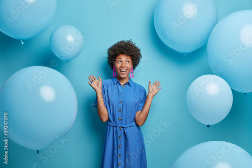 Positive carefree African American woman has festive mood raises hands and looks joyfully up ready for celebration dressed in festive clothing poses against blue background with big balloons