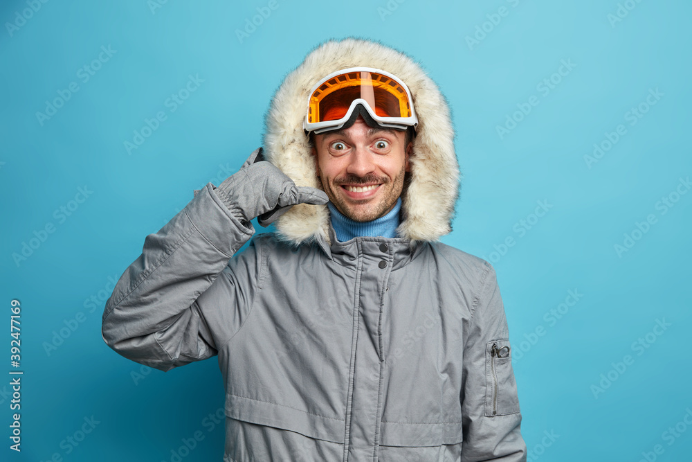 Happy male snowboarder wears winter jacket with hood makes phone gesture smiles positively isolated over blue background. Man skier enjoys favorite hobby asks to call him. Vacation and travel Happy male snowboarder wears winter jacket with hood makes phone gesture smiles positively isolated over blue background. Man skier enjoys favorite hobby asks to call him. Vacation and travel
