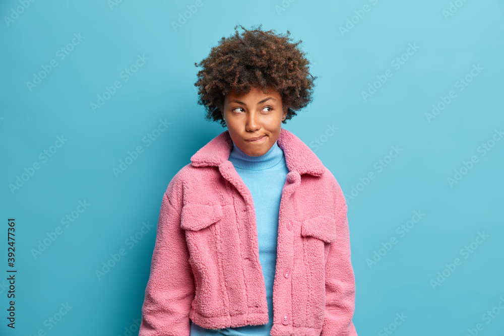 Photo of thoughtful dark skinned woman bites lips and looks away pensively has doubts and intention to do something as tries to remember something in mind wears fur coat isolated over blue wall. Photo of thoughtful dark skinned woman bites lips and looks away pensively has doubts and intention to do something as tries to remember something in mind wears fur coat isolated over blue wall.