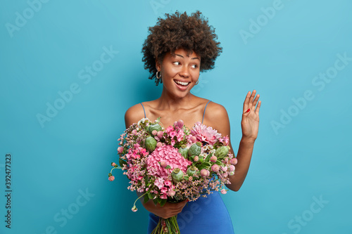 Positive dark skinned woman has curly hair waves palm and greets someone enjoys her birthday holds bouquet isolated over blue background. Feminine and floral concept. Lady with beautiful flowers