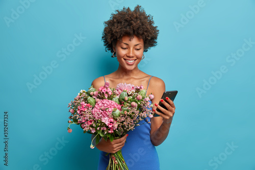 Happy smiling woman looks at smartphone display smiles broaldy holds mobile phone and receives messages of congratulation with her birthday holds big bunch of flowers isolated over blue studio wall