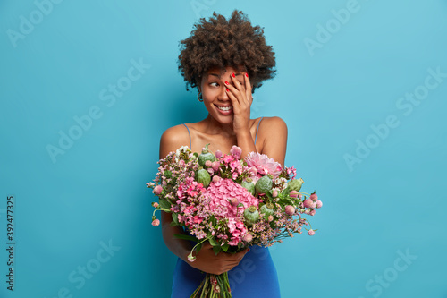 Photo of cheerful shy African American woman hides face with palm smiles happily wears dress holds big bouquet of flowers isolated over blue background. Pleased female receives present from husband