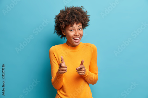 You got it. Happy young curly Afro American woman with curly hair points at camera with index fingers picks someone smiles broadly isolated over blue studio background. I choose you and order photo