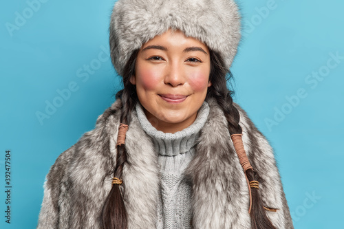 Photo of satisfied young siberia woman with two pigtails rosy cheeks smiles pleasantly at camera dresses for cold polar weather conditions isolated over blue background. Winter time concept. photo