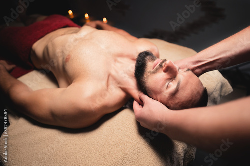 Male hands of a masseur doing neck massage to a bearded male athlete in a dark room of the massage room